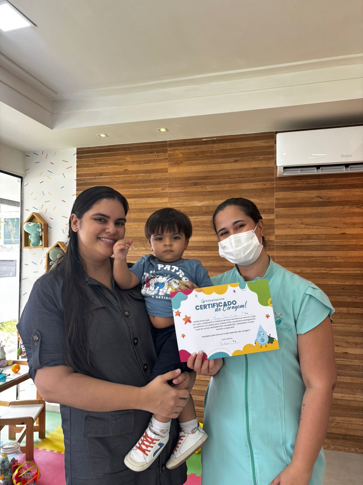 Mãe com o filho no colo ao lado de profissional da Prime Vaccine segurando o Certificado de Coragem