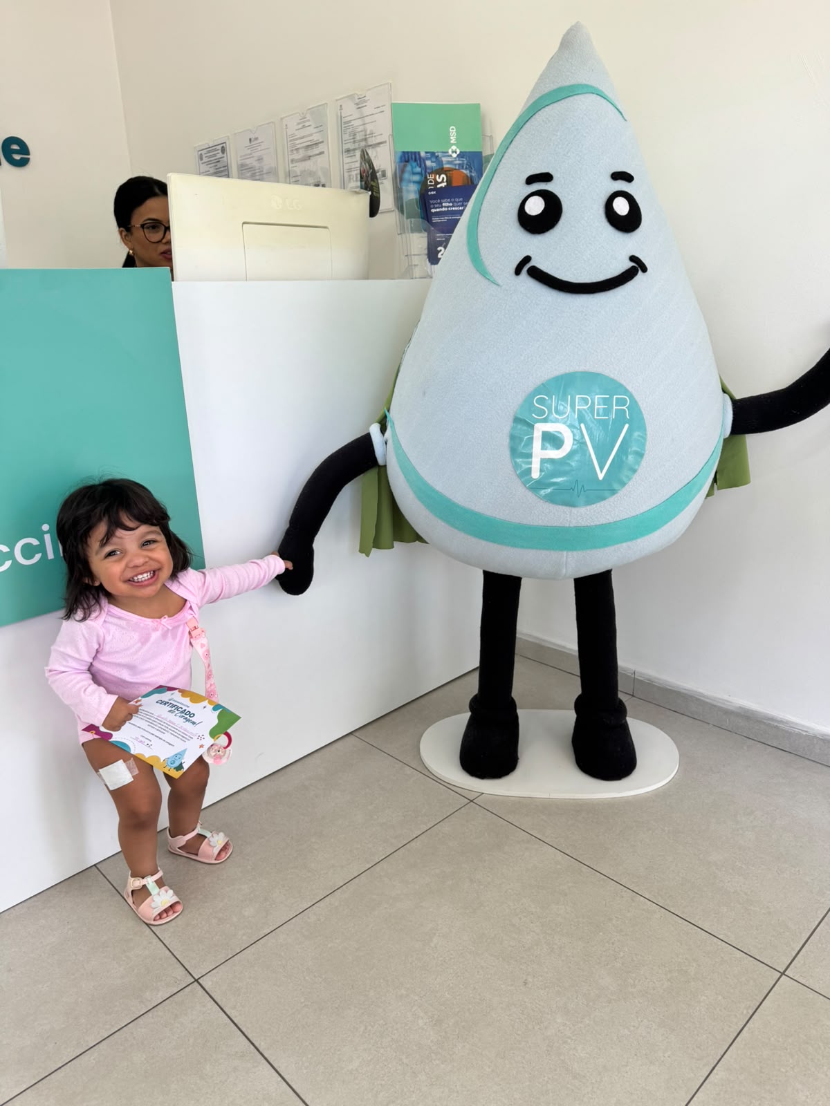 Criança sorrindo de mãos dadas com o mascote Super PV na recepção da Prime Vaccine