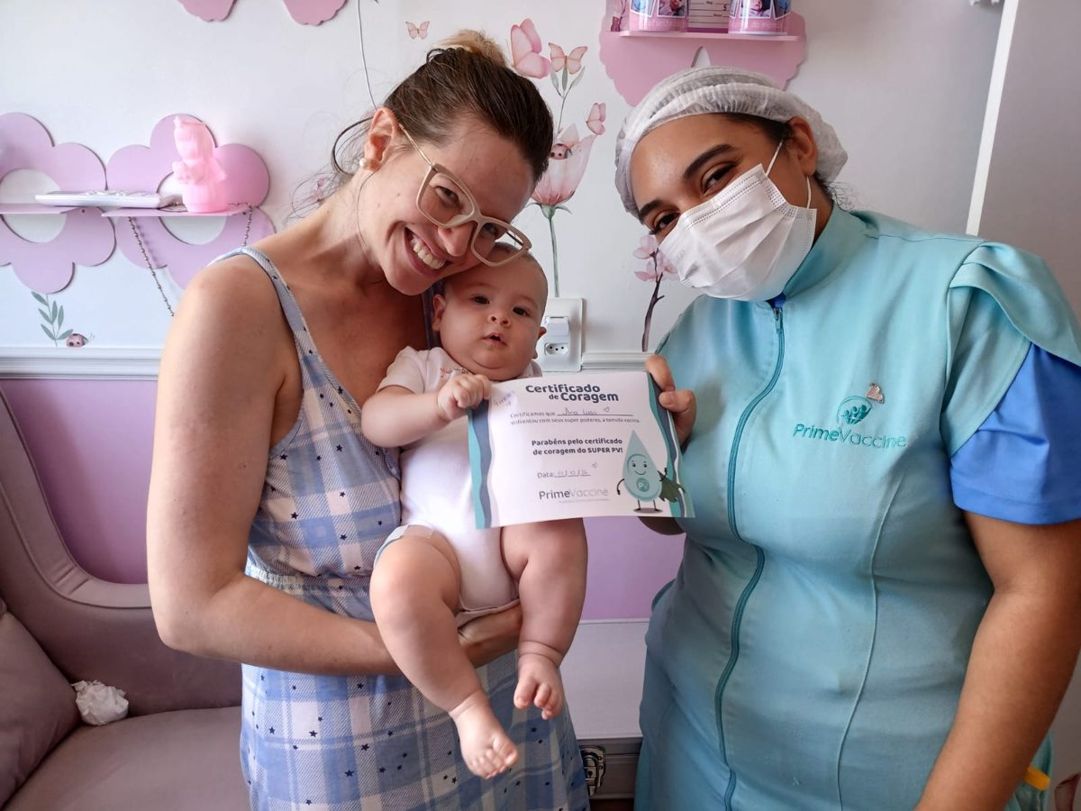 Mãe com bebê e profissional da Prime Vaccine segurando o Certificado de Coragem em casa