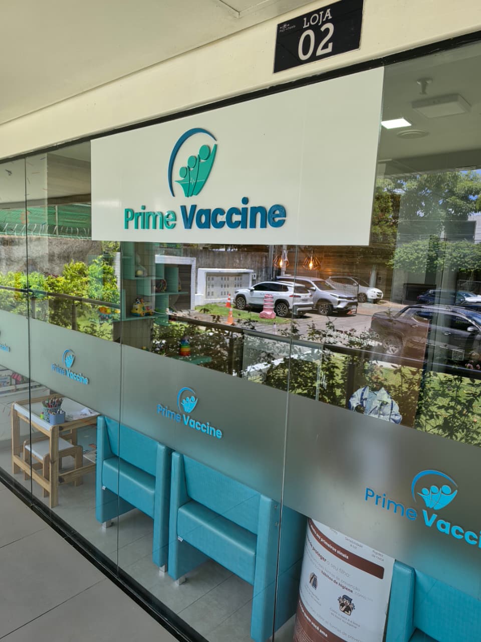 Fachada de vidro da Prime Vaccine na Loja 02, no Poço da Panela, com a logo da clínica e poltronas turquesa ao fundo